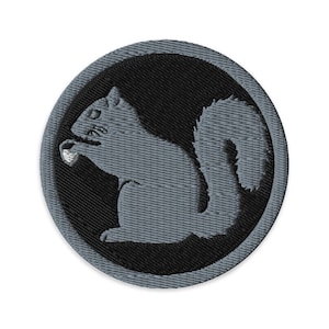 Op de afbeelding: Geborduurde patch met een grijze eekhoorn die een noot vasthoudt, tegen een zwarte cirkelvormige achtergrond. De eekhoorn is gedetailleerd met getextureerde steken en de patch heeft een grijze rand. Ideaal voor jassen, tassen of andere stoffen items.