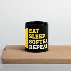 Peut inclure: Mug en céramique noire avec les mots "EAT SLEEP SOFTBALL REPEAT" en jaune et blanc. Un graphique de balle de softball jaune est sur le côté gauche de la tasse. La tasse est sur une surface en bois.