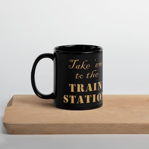 May include: Black ceramic coffee mug with a gold inscription that reads "Take 'em to the TRAIN STATION".