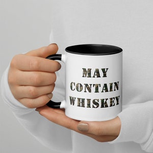 以下が含まれることがあります： 黒色の縁と取っ手を持つ白いセラミック製のマグカップ。マグカップには、黒い文字で「MAY CONTAIN WHISKEY」と書かれた迷彩柄のデザインが施されています。