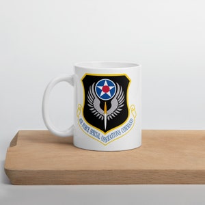 May include: White ceramic mug with a black and gold design. The design features a winged shield with a star and a dagger. The text "AIR FORCE SPECIAL OPERATIONS COMMAND" is written around the shield.