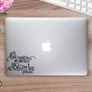 May include: A silver laptop with the Apple logo, featuring a black decal with the quote "To live would be an awfully big adventure - Peter Pan." A white mouse, keyboard, and pen are also visible.