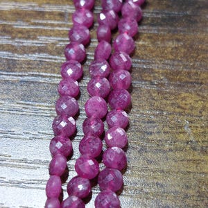 May include: Three strands of faceted, round, deep pink gemstones. The beads are strung on a pink cord, and the background is a dark wood grain.