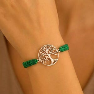 Puede incluir: Pulsera con dije de árbol de la vida plateado y banda verde tejida. El dije redondo presenta un diseño detallado de árbol. La pulsera se asegura con un cierre. Un accesorio elegante.