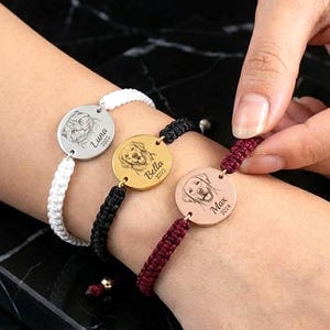 May include: Three personalized dog-themed bracelets. Each bracelet features a round metal charm with a dog portrait and a name and year. The bracelets have white, black, and burgundy braided cords. The bracelets are being worn on a wrist.