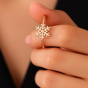 May include: A gold-colored ring featuring a snowflake design. The ring is held up by a person's fingers, showcasing the delicate details of the snowflake. The ring's band is thin and simple, complementing the snowflake charm.