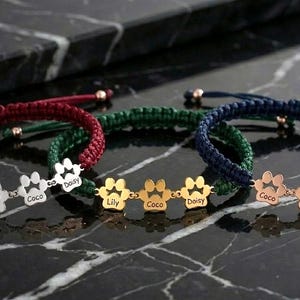 May include: Close-up of three personalized bracelets with paw print charms. The bracelets are red, green, and navy blue, with gold and silver-toned paw print charms. Each charm has a name engraved on it, such as "Lily", "Coco", and "Daisy".