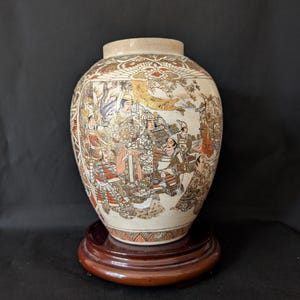 Puede incluir: Un jarrón de cerámica de color crema con una ilustración detallada y colorida de figuras con vestimenta tradicional japonesa. El jarrón se encuentra sobre un soporte redondo de madera. La obra de arte presenta detalles dorados, rojos y marrones.