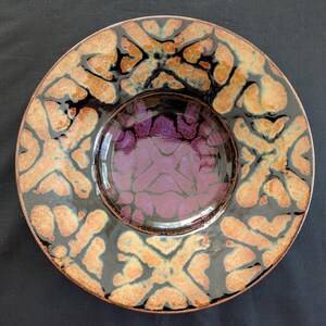 Puede incluir: Un plato de cerámica decorativo con un cuenco central. El plato presenta un patrón geométrico marrón, negro y naranja alrededor del borde. El cuenco tiene un diseño púrpura y rosa.