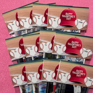 May include: A stack of Valentine's Day cards featuring baseball-themed designs. Each card displays a baseball heart with "TO" and "FROM" text, a red baseball cap with "HAPPY VALENTINE'S DAY!" printed on it, and a baseball field background.