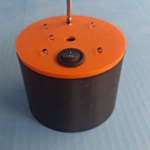 May include: A cylindrical device with a black base and an orange top. The top features a black on/off switch and several small, clear, dome-shaped elements. A metal rod extends from the center.