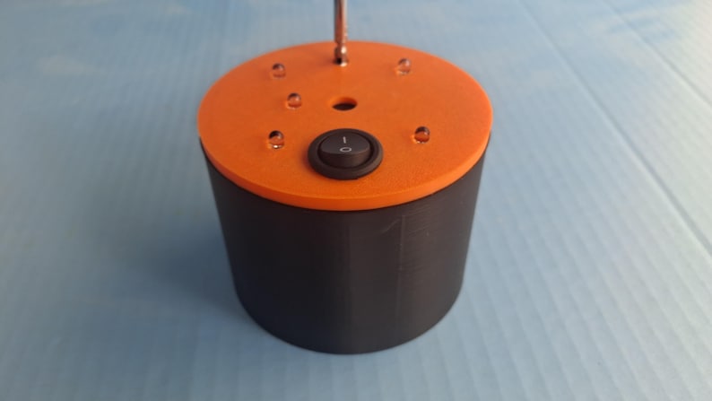 May include: A cylindrical device with a black base and an orange top. The top features a black on/off switch and several small, clear, dome-shaped elements. A metal rod extends from the center.