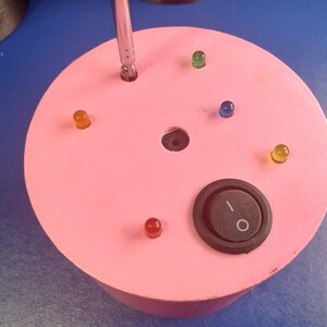 May include: A pink, circular electronic device with a black on/off switch and several colored LED lights. A screwdriver is inserted into a hole in the center. The device is on a blue surface.