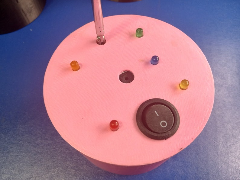 May include: A pink, circular electronic device with a black on/off switch and several colored LED lights. A screwdriver is inserted into a hole in the center. The device is on a blue surface.