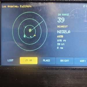 May include: A black electronic device with a rectangular screen displaying a radar interface. The screen shows Los Angeles, California, 2:12 PM, IN RANGE 39, NEAREST N232LA, A550, 975 ft, 39 kt, 2 nm, and buttons labeled LIST, 25 NM, PLACE, BRIGHT, WIFI.