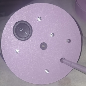 May include: A close-up of a light purple, circular object with a black on/off switch and several small, clear, round embellishments. A thin, light purple rod extends from the side. The object appears to be a lamp base.