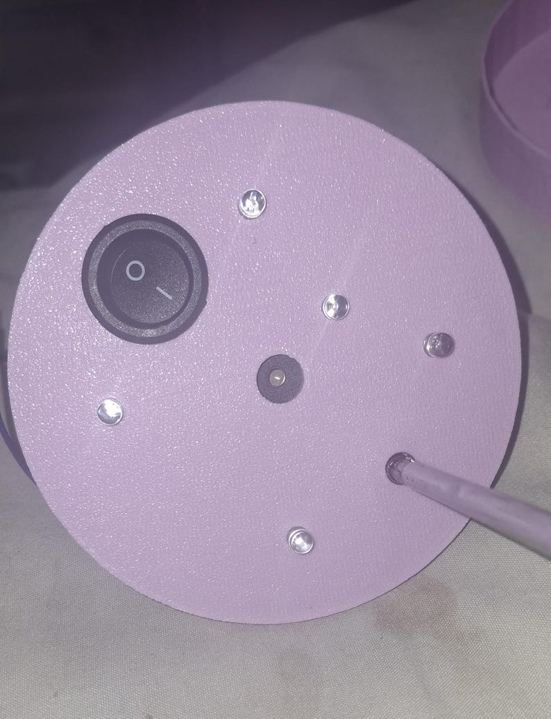 May include: A close-up of a light purple, circular object with a black on/off switch and several small, clear, round embellishments. A thin, light purple rod extends from the side. The object appears to be a lamp base.