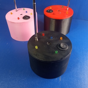May include: Three cylindrical devices in pink, black, and red, each with a silver antenna, colored LED lights, and an on/off switch. The devices are on a blue surface.
