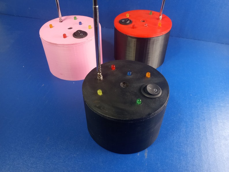 May include: Three cylindrical devices in pink, black, and red, each with a silver antenna, colored LED lights, and an on/off switch. The devices are on a blue surface.