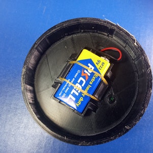May include: A black, circular 3D-printed container with a 9-volt battery and a blue and yellow battery. The battery is secured with a rubber band. The container sits on a blue surface.