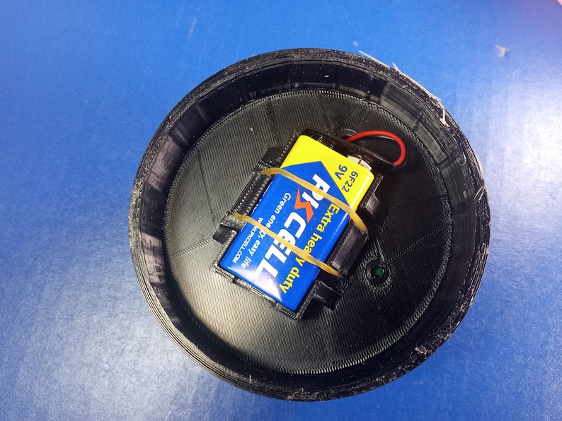 May include: A black, circular 3D-printed container with a 9-volt battery and a blue and yellow battery. The battery is secured with a rubber band. The container sits on a blue surface.