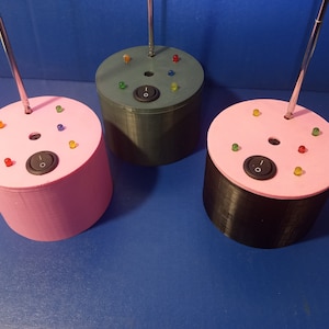 May include: Three cylindrical devices with a pink, green, and black base. Each has a circular top with colored LED lights, a black on/off switch, and a silver antenna. The devices are on a blue surface.