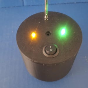 May include: A cylindrical black electronic device featuring a single rocker switch, a small metallic antenna extending from the top, and two illuminated indicator lights, one glowing amber and the other glowing green, set against a blue background.