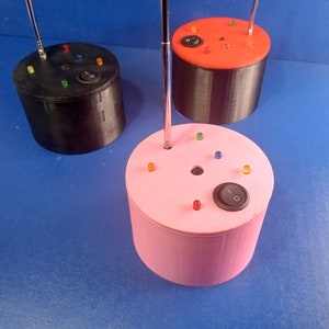 May include: Three cylindrical electronic devices in black, red, and pink. Each has a silver antenna, colored LED lights, and an on/off switch. The devices are on a blue surface.