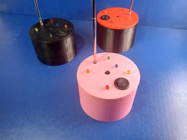 May include: Three cylindrical electronic devices in black, red, and pink. Each has a silver antenna, colored LED lights, and an on/off switch. The devices are on a blue surface.