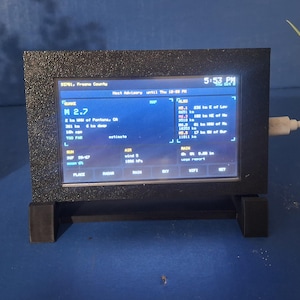 May include: A black rectangular digital weather display device with a textured frame, mounted on a small stand. The screen shows blue text and data, including time, location, and meteorological information, with a white power cable attached to the side.