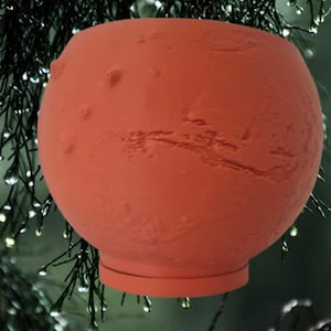 May include: A spherical terracotta-colored planter featuring a textured surface that mimics the cratered and uneven topography of a planet, with a flat circular base and an open top, suspended against a backdrop of dark green pine needles.