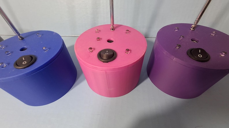 May include: Three cylindrical, colorful objects with a switch and a metal rod extending from the top. The objects are blue, pink, and purple. Each has a black on/off switch and small clear accents.