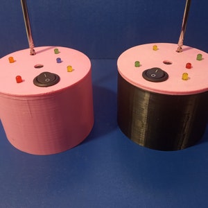 May include: Two cylindrical devices, one pink and one black, with a pink top. Each has a silver antenna, a black on/off switch, and several colored LED lights. The devices are on a blue surface.