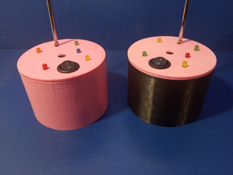 May include: Two cylindrical devices, one pink and one black, with a pink top. Each has a silver antenna, a black on/off switch, and several colored LED lights. The devices are on a blue surface.