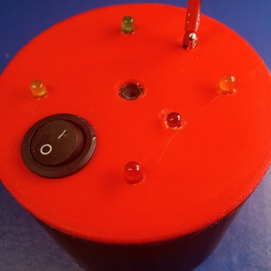 May include: A red electronic device with a black toggle switch and several small LED lights. The lights are various colors, including red, yellow, and green. The device has a cylindrical shape.