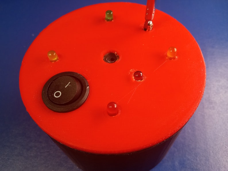 May include: A red electronic device with a black toggle switch and several small LED lights. The lights are various colors, including red, yellow, and green. The device has a cylindrical shape.