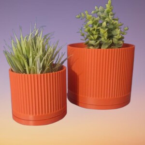 May include: Two cylindrical orange planters with a vertical ribbed texture. Each planter features a matching base tray and contains a green artificial plant. The smaller planter is positioned in the front left, while the larger one is behind it.