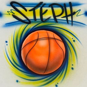 Puede incluir: Una pelota de baloncesto de color naranja y negro est&aacute; rodeada por un dise&ntilde;o de espiral azul y amarillo. La palabra "STEPH" est&aacute; escrita en negro sobre la pelota.