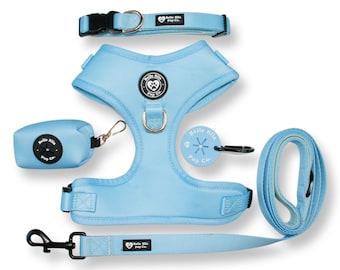 May include: A light blue dog harness, leash, collar, and poop bag holder set. The set features a black buckle on the collar and a black clip on the leash. The harness has a black metal ring and a black plastic buckle. The poop bag holder has a black circle with a paw print in the center. The set is branded with the text "Belle Ella Pup Co."