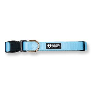 Peut inclure: Un collier pour chien bleu clair avec une boucle noire et un anneau en D en m&eacute;tal dor&eacute;. Le collier a une &eacute;tiquette noire avec du texte blanc qui dit "Belle Ella Pup Co."
