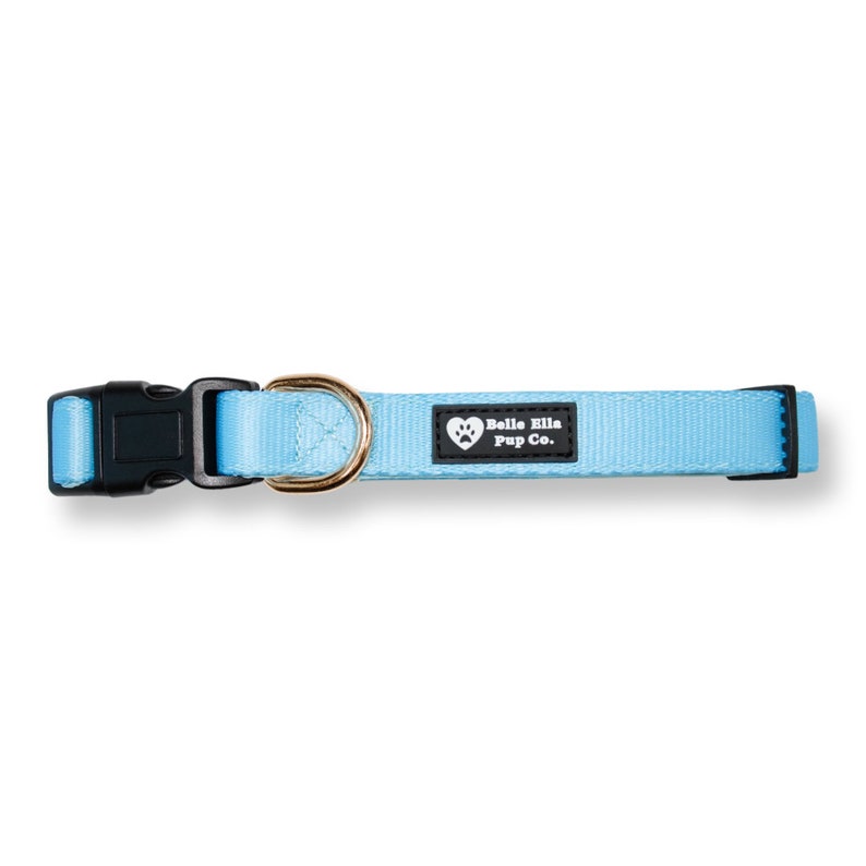 Peut inclure: Un collier pour chien bleu clair avec une boucle noire et un anneau en D en m&eacute;tal dor&eacute;. Le collier a une &eacute;tiquette noire avec du texte blanc qui dit "Belle Ella Pup Co."
