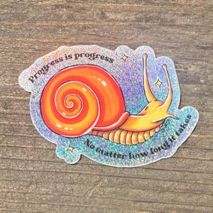 Puede incluir: Pegatina holográfica con un caracol de dibujos animados con concha naranja y cuerpo amarillo. La pegatina tiene el texto "Progress is progress, no matter how long it takes" en un diseño circular. La pegatina tiene un acabado brillante e iridiscente.