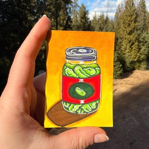 Puede incluir: Una pintura de acuarela de un tarro de pepinillos. El tarro es verde con una etiqueta roja y un gráfico de pepinillo verde. El fondo es amarillo y el tarro se asienta sobre una superficie marrón. La pintura se sostiene en una mano.
