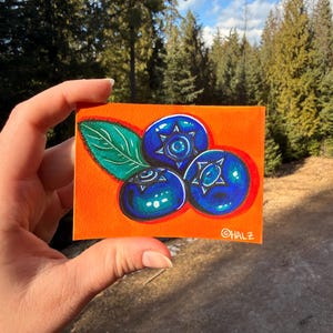 Puede incluir: Una obra de arte pintada a mano que representa tres arándanos con detalles en forma de estrella y una hoja verde sobre un fondo naranja. Los arándanos son de un azul vibrante con acentos azul oscuro. La obra está firmada "©HALZ".