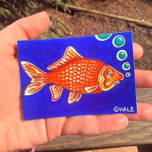 Puede incluir: Ilustración de un pez dorado naranja brillante sobre un fondo azul, con burbujas y la firma del artista. El pez tiene escamas y aletas detalladas, sobre un fondo de líneas acuáticas. La obra de arte se sostiene en una mano.