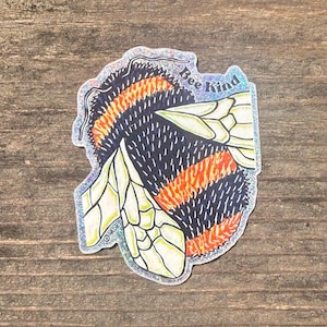 Puede incluir: Una pegatina holográfica con una abeja estilizada con detalles en negro, naranja y blanco. El cuerpo de la abeja tiene rayas naranjas y una textura negra similar a la piel. El texto "Bee Kind" está escrito en la esquina superior derecha.