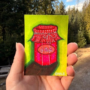 Puede incluir: Pintura a la acuarela de un tarro de mermelada con tapa a cuadros rojos y blancos y una etiqueta que dice "JAM". El tarro es rosa con lunares blancos, sobre un fondo verde.