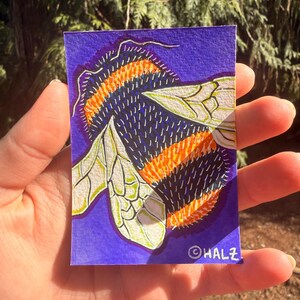 Puede incluir: Una obra de arte rectangular, sostenida en la mano, que presenta una ilustración detallada de una abeja. El cuerpo de la abeja está rayado con naranja y negro, con alas blancas y verdes sobre un fondo púrpura. La firma del artista, "©HALZ", es visible.