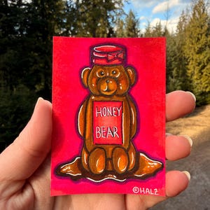 Puede incluir: Ilustración dibujada a mano de un tarro de miel con forma de oso sobre un fondo rojo. El tarro marrón con forma de oso tiene una tapa roja con un lazo y las palabras "HONEY BEAR" en la parte delantera. La firma del artista, "©HALZ", está en la esquina inferior derecha.