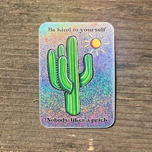 Puede incluir: Una pegatina rectangular con un fondo holográfico que presenta un cactus verde y un sol amarillo. El texto "Be kind to yourself" está en la parte superior y "Nobody likes a prick" en la inferior.